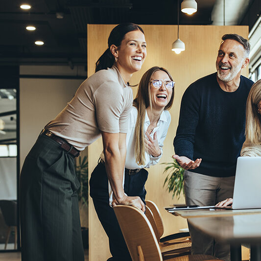 Happy businesspeople laughing while collaborating on a new project in an office. Group of diverse businesspeople using a laptop while working together in a modern workspace.